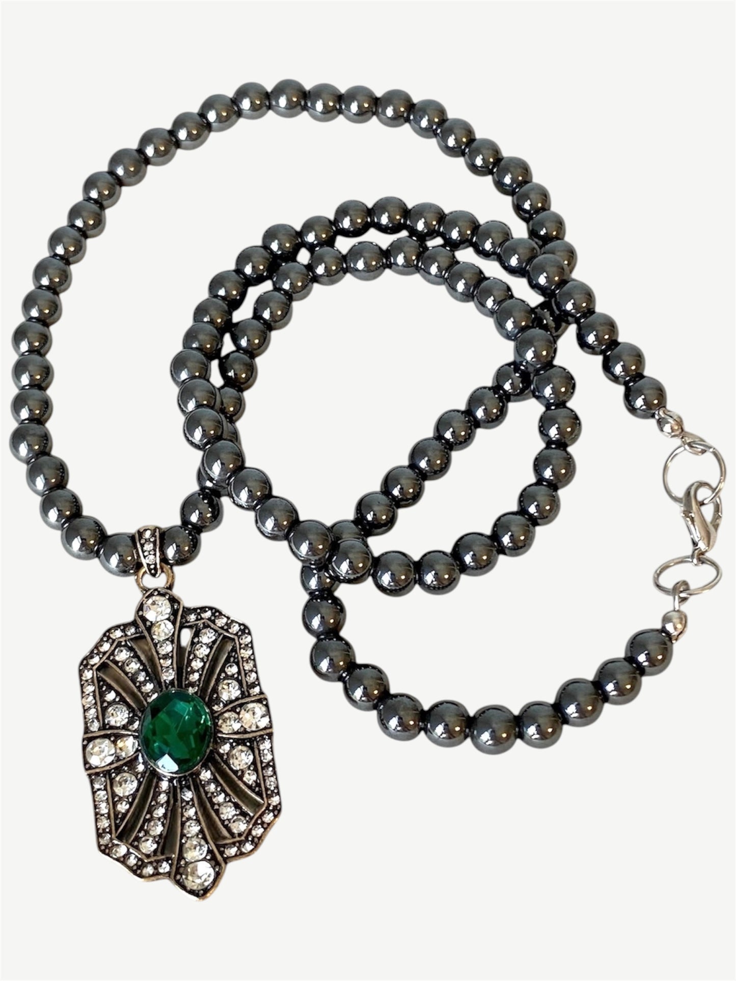 A dark gray beaded necklace with a vintage Art Deco style pendant featuring a green emerald center stone and clear rhinestone accents, displayed on a light beige  background.