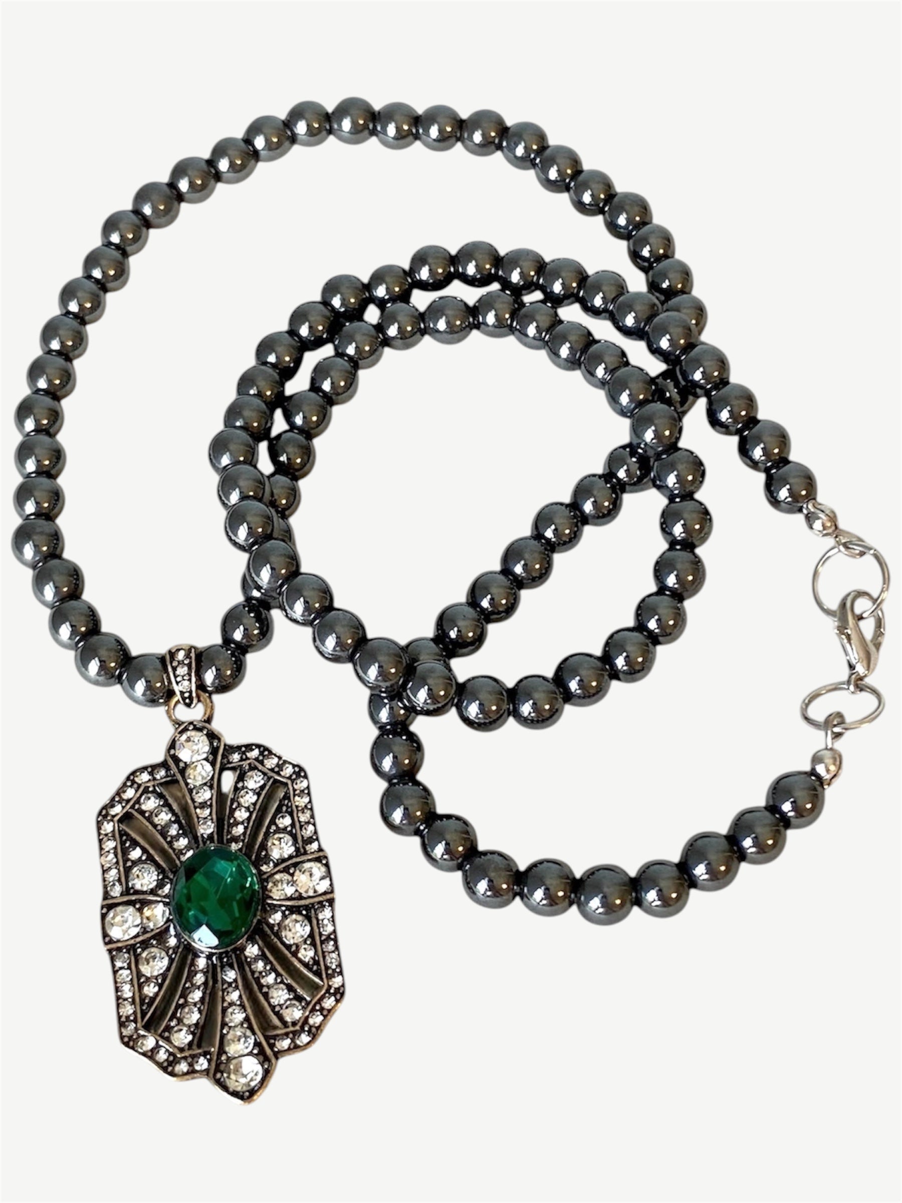 A dark gray beaded necklace with a vintage Art Deco style pendant featuring a green emerald center stone and clear rhinestone accents, displayed on a light beige  background.