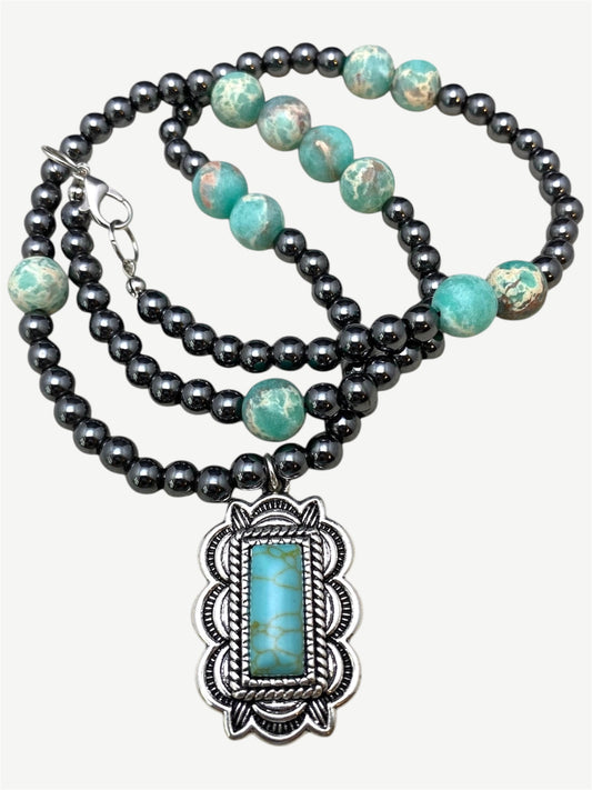 A bohemian-style double-strand necklace featuring dark gray beads and turquoise-colored beads, with a large silver-toned pendant containing a rectangular turquoise stone.