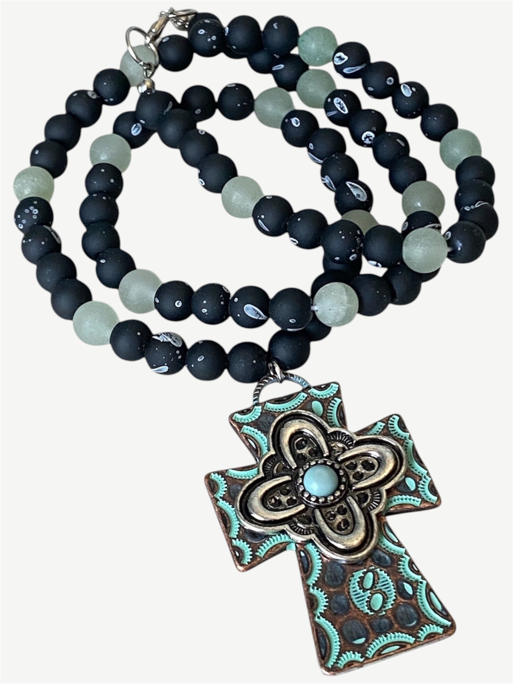 A beaded necklace with matte black and pale green beads, featuring a large ornate cross pendant with turquoise and bronze detailing, shown on a light beige background.