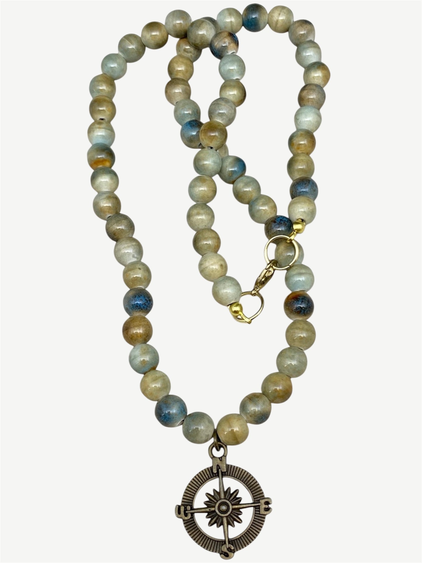 A beaded necklace featuring ceramic beads in earthy brown, beige, and blue-gray tones, with a brass compass pendant and gold-tone clasp, shown on a light beige background.