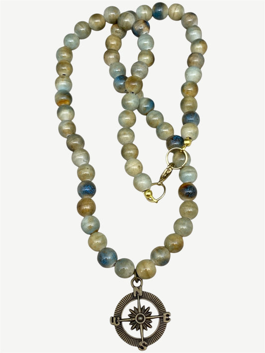 A beaded necklace featuring ceramic beads in earthy brown, beige, and blue-gray tones, with a brass compass pendant and gold-tone clasp, shown on a light beige background.