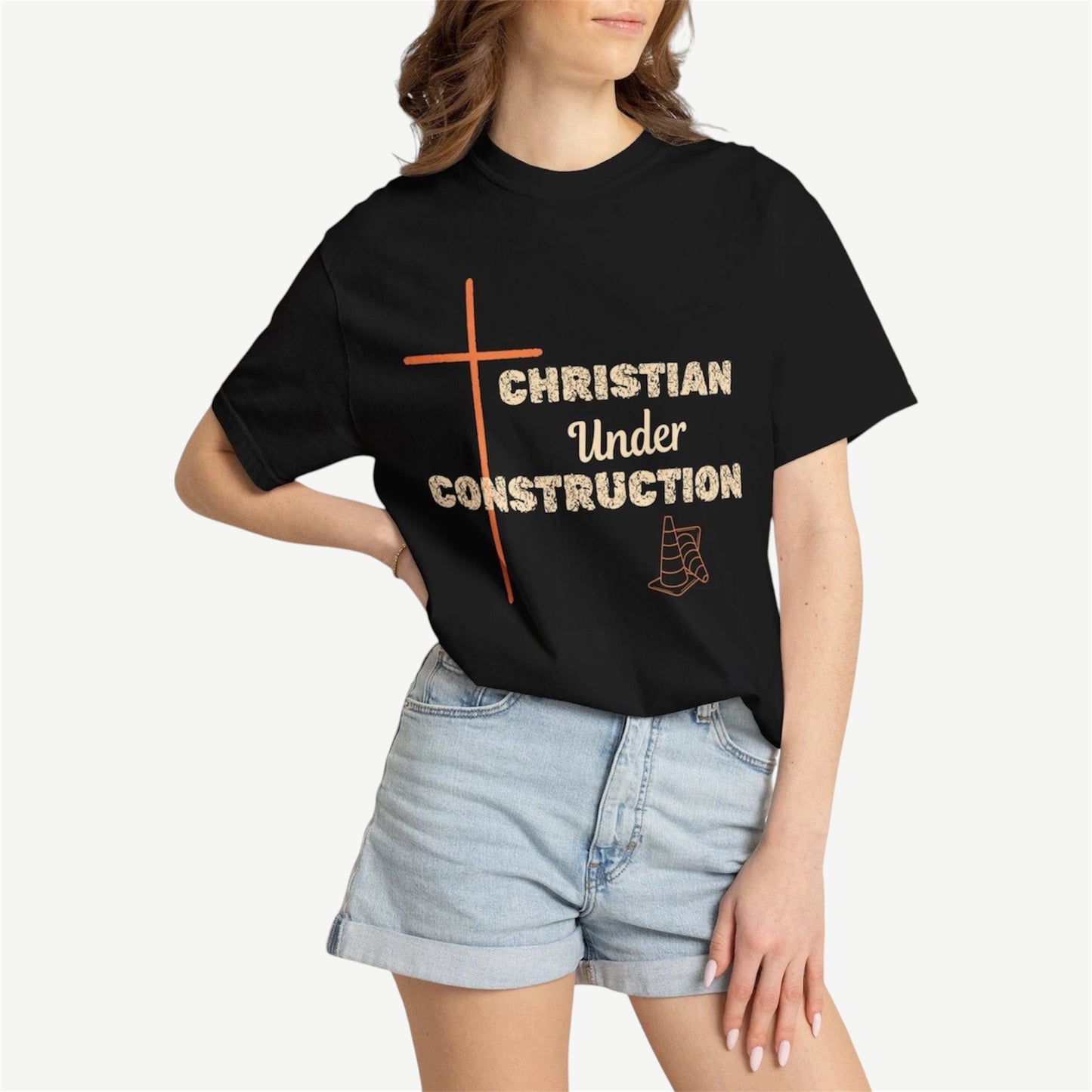 A woman wearing a black graphic t-shirt with a orange cross and the text 'CHRISTIAN Under CONSTRUCTION' in beige, paired with light-wash denim shorts, against a light beige background.