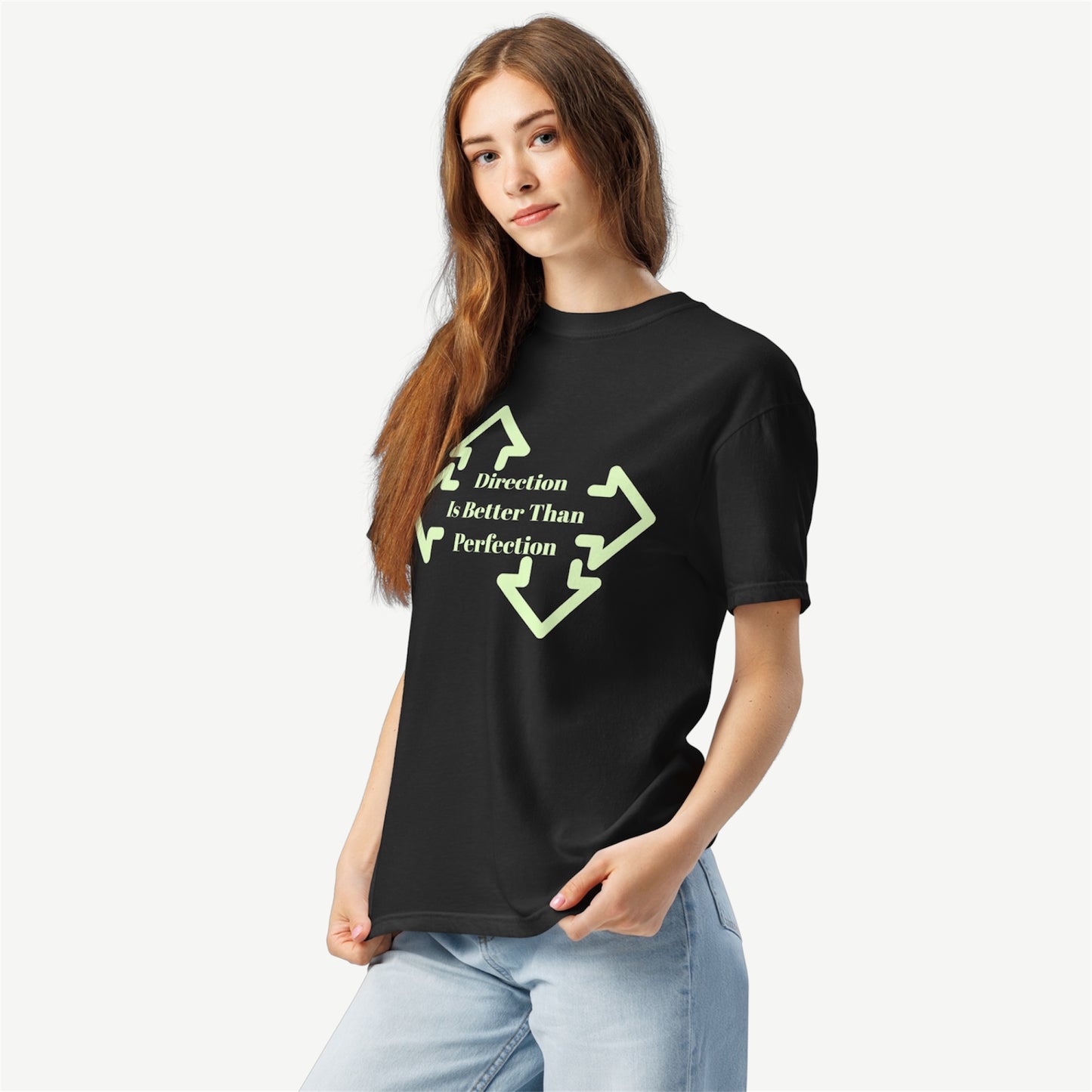 A woman modeling a black short-sleeve t-shirt with a light green graphic that reads 'Direction is Better Than Perfection' inside a broken arrowhead design, paired with light blue jeans against a light gray background.